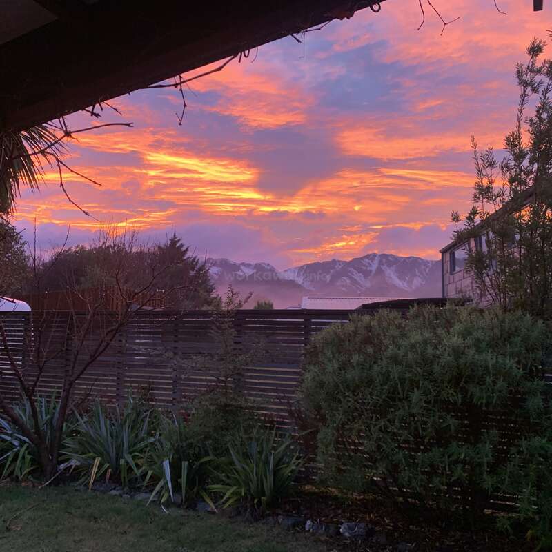Una espectacular puesta de sol pinta el cielo con tonos naranjas y morados sobre montañas nevadas, un jardín y una valla de madera. Árboles en silueta enmarcan la apacible vista.