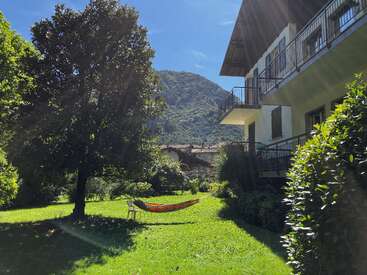 A peaceful green garden with a hammock and chair in the sun, surrounded by trees, a building, and a scenic mountain under a clear blue sky.