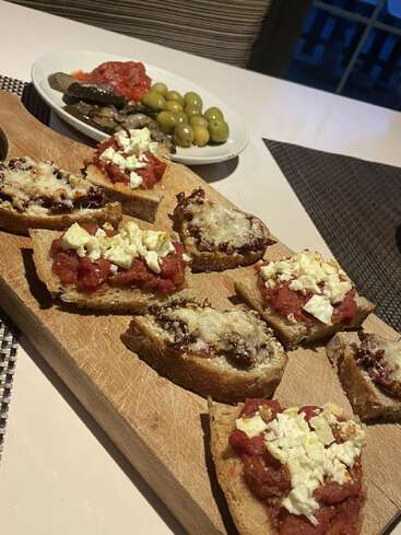 Esta imagem mostra uma tábua de madeira com fatias de pão torrado cobertas com tomate, queijo e molho. Ao fundo, há um prato com azeitonas e legumes.
