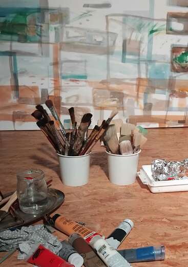 Dois copos brancos cheios de pincéis estão sobre uma mesa de madeira. Tubos de tinta espalhados, uma jarra de água e obras de arte abstratas completam essa cena criativa e artística.