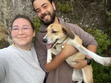 A smiling woman and man pose outdoors with a happy brown and white dog. They appear joyful, surrounded by greenery and rocks, creating a warm, friendly atmosphere.
