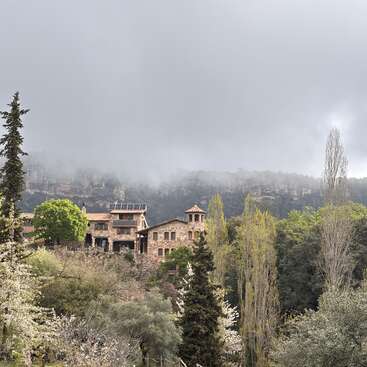 A rustic stone mansion sits nestled among lush trees and blooming flowers. Mist and fog envelop the mountainous background, creating a tranquil, mysterious atmosphere.