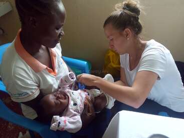 Eine Frau hält ein Baby im Arm, während eine andere Frau dem Baby beim Anziehen hilft. Sie sitzt auf einem blauen Stuhl in einem Raum mit beiger Wand.