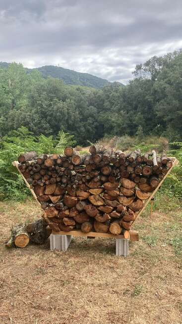 A neatly stacked pile of firewood rests on a homemade wooden stand outdoors, surrounded by green foliage, with forested hills and cloudy skies in the background.