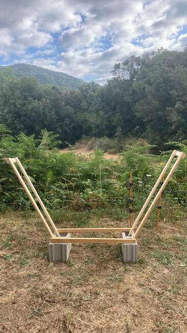 A simple wooden frame structure rests on concrete blocks in a grassy clearing, surrounded by lush greenery and hills, under a partly cloudy sky.