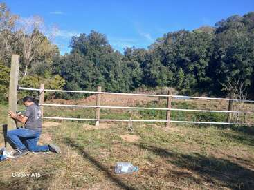 A man kneels on grass, working on a wooden fence post. Trees and clear blue sky are in the background; a plastic bottle lies nearby.