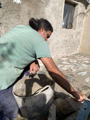 A man kneels outside near a stone wall, mixing and applying cement from a bucket, working carefully on constructing or repairing a stone surface in sunlight.