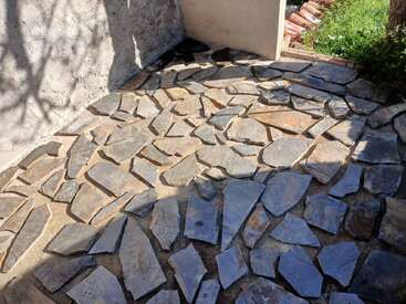 This image shows a patio or pathway made of irregularly shaped flat stones arranged closely together, bordered by walls and greenery, with sunlight casting distinct shadows.