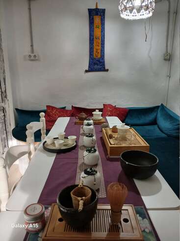 A cozy tea room setup with a white table, traditional Chinese tea set, colorful cushions, and wall hanging. Warm, inviting atmosphere for tea ceremonies or gatherings.