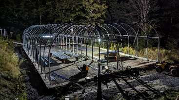 A steel-frame structure is under construction at night, illuminated by work lights. Materials and equipment are scattered around the site, surrounded by trees and vegetation.