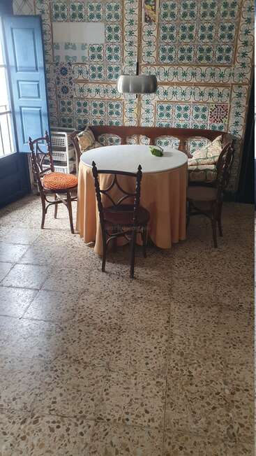 Une salle à manger de style rétro avec une table ronde, une nappe orange, des chaises en bois, un banc avec des coussins, des carreaux muraux aux motifs élaborés, un sol en terrazzo et des volets bleus.