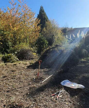 Une scène de jardin baignée de soleil montre des outils de jardinage, une pelle et un sac posés sur un sol sec. De grands arbres et des buissons entourent l'endroit sous un ciel dégagé.