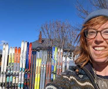 Une personne souriante, portant des lunettes et un bonnet bleu, se tient dehors par une journée ensoleillée, avec une rangée de skis colorés posés à la verticale derrière elle, près d'une maison.