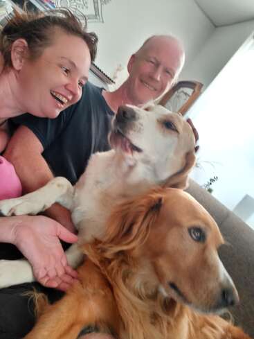 Ein fröhlicher Moment, den zwei lächelnde Menschen und ihre beiden Hunde auf einem Sofa teilen und der eine Atmosphäre von Glück, Wärme und liebevoller Zweisamkeit zu Hause einfängt.