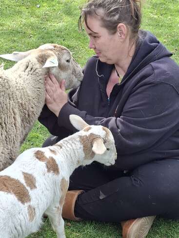 Eine Frau sitzt im Gras, hält sanft das Gesicht eines Schafs in den Händen und schaut es liebevoll an. Ein junges Zicklein mit braunen Flecken steht daneben und sorgt für einen friedlichen, herzlichen Moment auf dem Bauernhof.