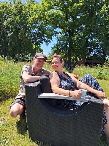 Ein lächelndes Paar entspannt sich an einem sonnigen Tag im Freien. Die Frau sitzt mit einer Tasse in einem Korbstuhl, umgeben von grünem Gras und hohen, belaubten Bäumen.