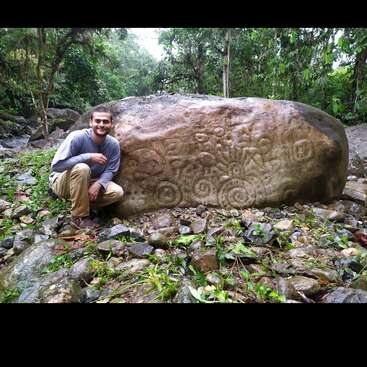 A imagem mostra uma pedra grande com um desenho gravado, com uma linha vertical proeminente e uma linha horizontal na parte inferior, em um cenário natural ao ar livre.
