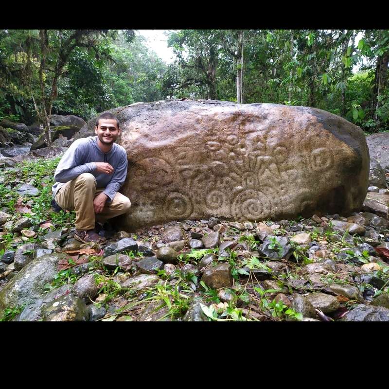 Das Bild zeigt einen großen Felsen mit einem eingravierten Design mit einer markanten vertikalen Linie und einer horizontalen Linie am unteren Rand vor einer natürlichen Kulisse.