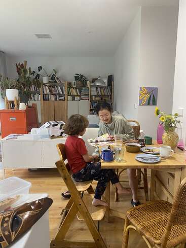 A woman and child sit at a wooden table, sharing breakfast and reading. The room is cozy, with bookshelves, plants, and a vase of flowers.