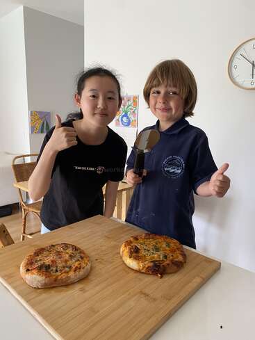 Two children stand by a table with two homemade pizzas, smiling and giving thumbs up. One holds a pizza cutter; the atmosphere is cheerful and proud.