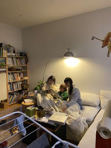 Three people sit cozily on a white couch under a blanket, reading together. Warm lighting, bookshelves, tea, and indoor plants create a relaxing, homey atmosphere.