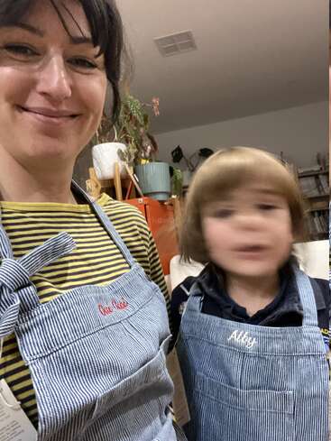 A woman and a child, both wearing striped aprons, smile indoors. The child is slightly blurred, and houseplants are visible in the cozy background.