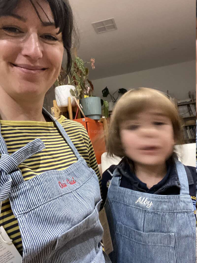 A woman and a child, both wearing striped aprons, smile indoors. The child is slightly blurred, and houseplants are visible in the cozy background.