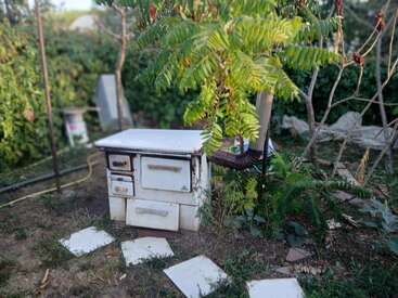 Un vieux poêle rouillé est abandonné à l'extérieur, au milieu de l'herbe, de carreaux cassés et de plantes envahissantes. Un petit arbre pousse à côté, masquant partiellement le poêle.