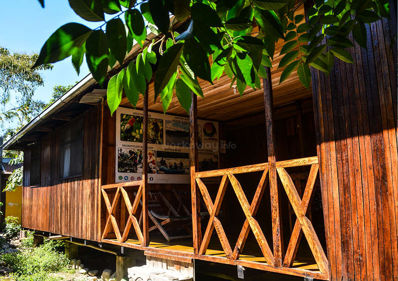 La imagen representa un edificio de madera con un porche, provisto de una barandilla y un letrero, rodeado de árboles y un cielo azul despejado en un día soleado.