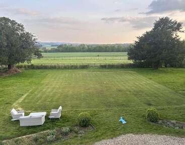 A peaceful, large green lawn with scattered outdoor furniture, surrounded by fields and trees. Distant hills, a soft sunset sky, and a blue toy are visible.