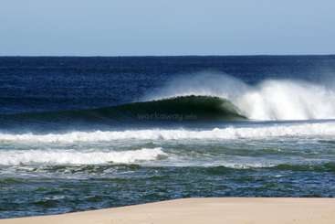 L'image représente une scène de plage sereine avec une grande étendue d'eau, une vague proéminente et un rivage sablonneux, évoquant un sentiment de tranquillité et de beauté naturelle.
