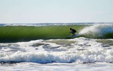 L'image représente une personne surfant sur une vague, avec l'océan et le ciel visibles en arrière-plan. La personne est au centre du cadre, entourée d'eau.
