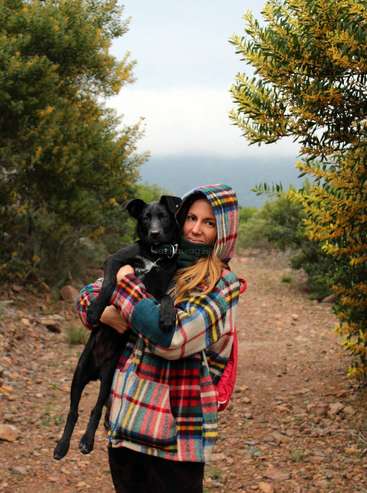 Une femme vêtue d'une veste à capuche à carreaux tient un chien noir sur un chemin de terre entouré de buissons et d'arbres, sous un ciel couvert.