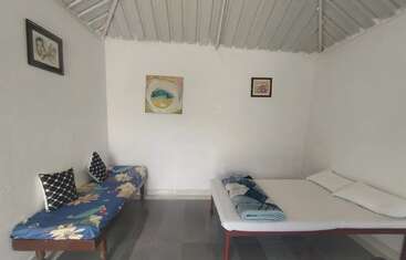 Auf dem Bild sieht man ein einfaches Zimmer mit einem Bett, einem Sofa mit Blumenmuster, drei gerahmten Bildern an weißen Wänden, Fliesenboden und einem schrägen Metalldach.