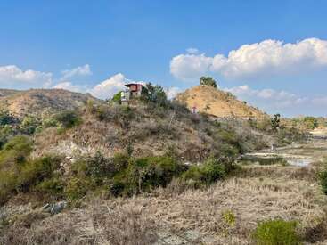 Ein kleines Haus steht auf einem trockenen, grasbewachsenen Hügel, umgeben von spärlicher Vegetation, unter einem strahlend blauen Himmel mit flauschigen weißen Wolken und fernen Hügeln.