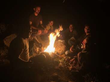 Sechs Leute sitzen nachts um ein Lagerfeuer herum und genießen die Wärme und das Licht. Ihre Gesichter strahlen vor Glück, während sie Geschichten austauschen und in der Natur zusammenkommen.