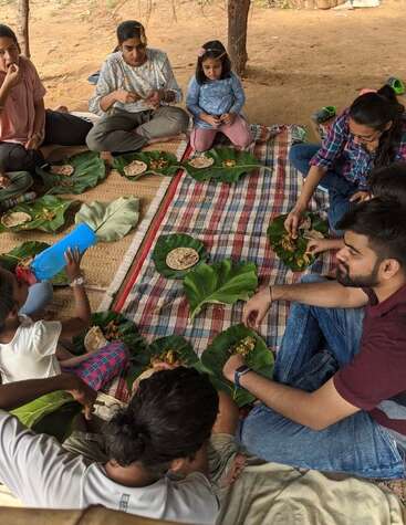 Eine Gruppe von Leuten, darunter auch Kinder, sitzt draußen auf einer Matte, teilt sich eine Mahlzeit, die auf großen grünen Blättern serviert wird, und genießt gemeinsam traditionelles Essen und Gespräche.