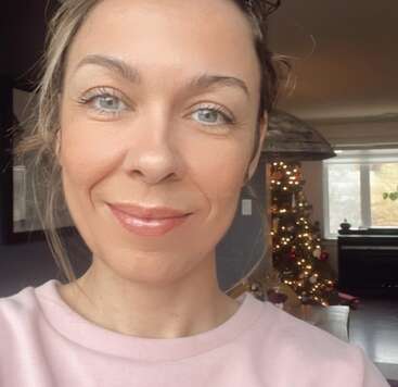 Une femme aux yeux bleus sourit légèrement, vêtue d'un haut rose pâle. Derrière elle, un sapin de Noël décoré et illuminé trône dans un salon confortable et ensoleillé.