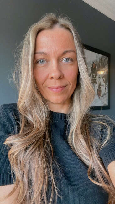 Une femme aux longs cheveux bruns ondulés et aux yeux bleus porte un haut noir et sourit légèrement. Une photo encadrée est accrochée au mur sombre derrière elle.
