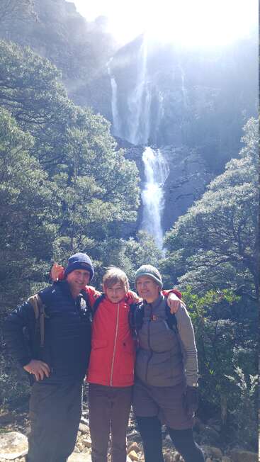 A family of three, warmly dressed, smiles in front of a tall, sunlit waterfall surrounded by lush green trees during a scenic outdoor hike.