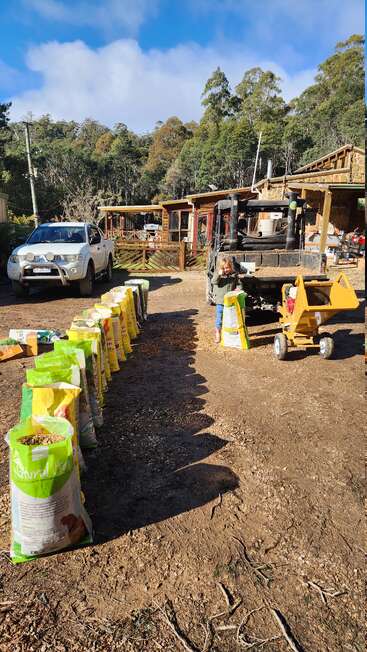 A child stands by a utility vehicle and wood chipper, with bags of animal feed lined up on a dirt driveway, surrounded by forest and rustic buildings.