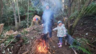 A woman and child, dressed warmly, stand by a small campfire in a forest. Smoke rises. Green ferns, trees, and a dog surround them.