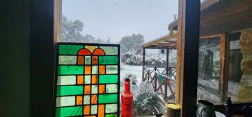 A snowy landscape is visible through a window with a colorful stained-glass panel. Wooden porch, trees, and decorative bottles create a cozy, inviting indoor scene.