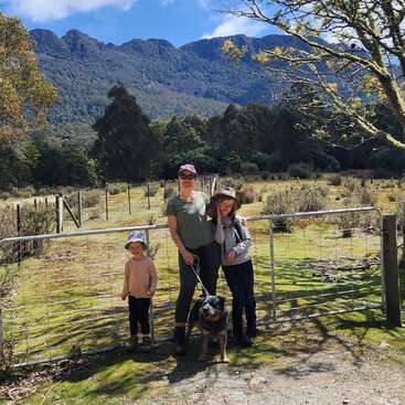 Three people and a dog stand by a metal gate in a picturesque, sunny countryside with trees, grassy field, and mountains in the background, smiling for the camera.