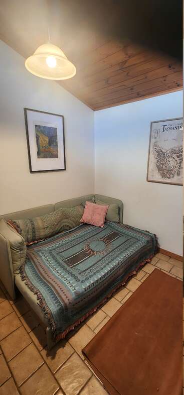 A cozy corner with a green couch, patterned blankets, and pillows below framed wall art. Warm lighting, tiled floor, and a wooden ceiling create a snug atmosphere.
