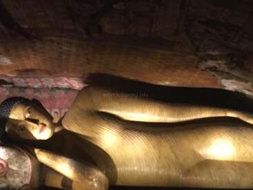Cette image montre une grande statue de Bouddha couché, baignée d'une lumière chaleureuse, à l'intérieur d'une grotte faiblement éclairée, dont les murs sont ornés de motifs complexes et de décorations peintes.