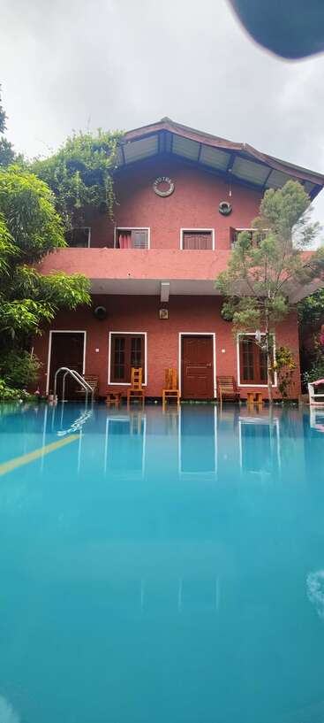 Un hôtel rouge et chaleureux doté d'un balcon, niché au cœur d'une végétation luxuriante. On y voit des chaises en bois et, au premier plan, une piscine aux eaux d'un bleu limpide.