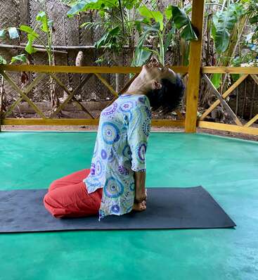 Une personne pratique le yoga sur un tapis dans un cadre extérieur paisible, à genoux et le dos cambré, entourée de plantes vertes et d'une clôture en bois. Ambiance sereine.