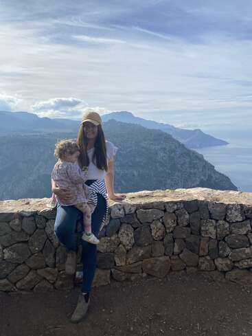 Das Bild zeigt eine Frau und ein Kind, die für ein Foto auf einer Steinmauer mit Blick auf eine bergige Landschaft und ein Gewässer unter einem bewölkten Himmel posieren.
