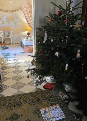 Un sapin de Noël décoré se trouve près d'une porte. Au-delà, une salle de jeux confortable pour les enfants avec des jouets, une carte, un lit et un éclairage doux crée une atmosphère chaleureuse et festive.